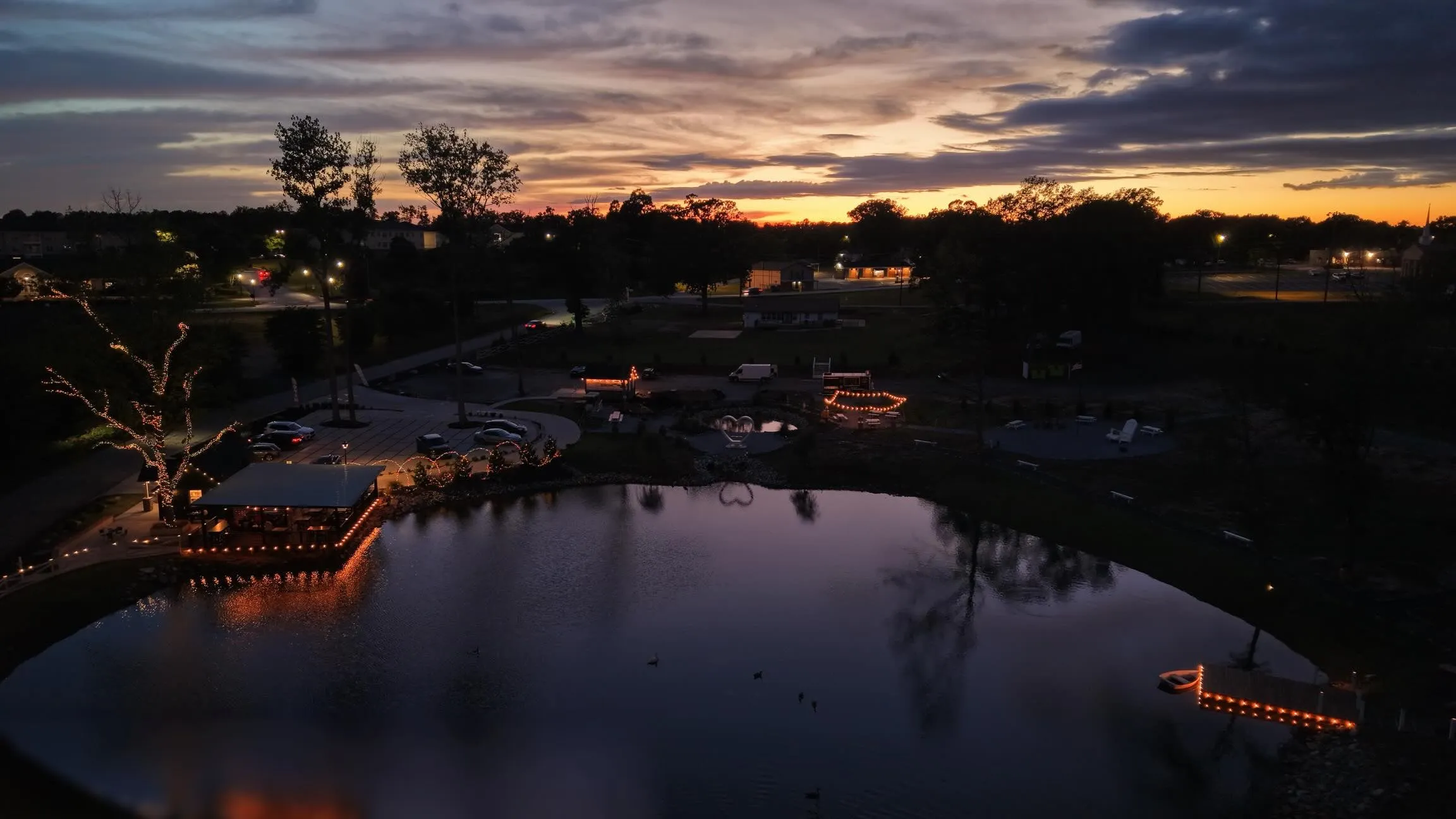 Drone view of the pond after sunset with the pavilion, dock and paths lit by string lights