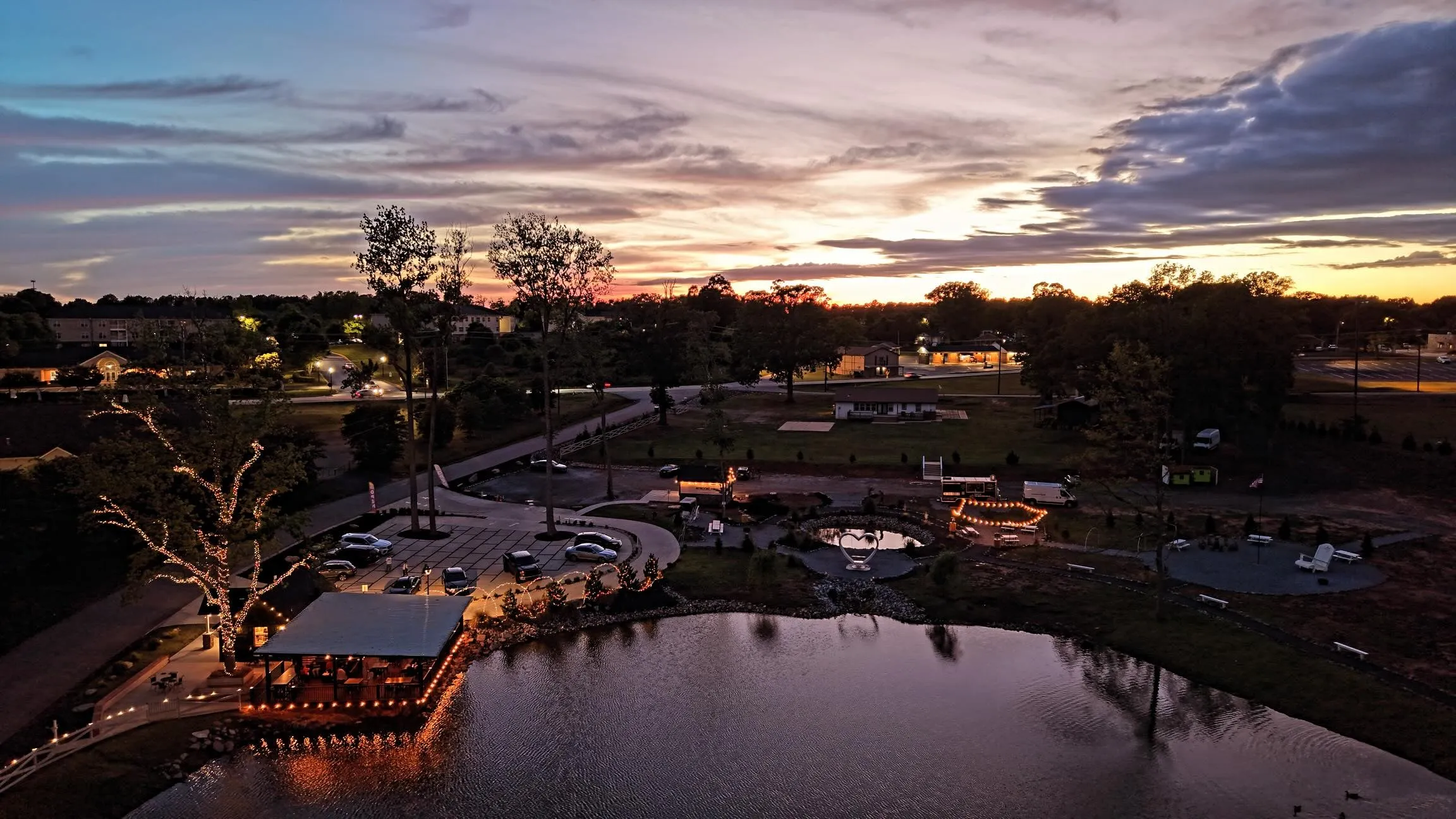 Drone view of 121 Coffee at sunset: the lit pavilion on the pond, the parking circle, and string lights across the grounds