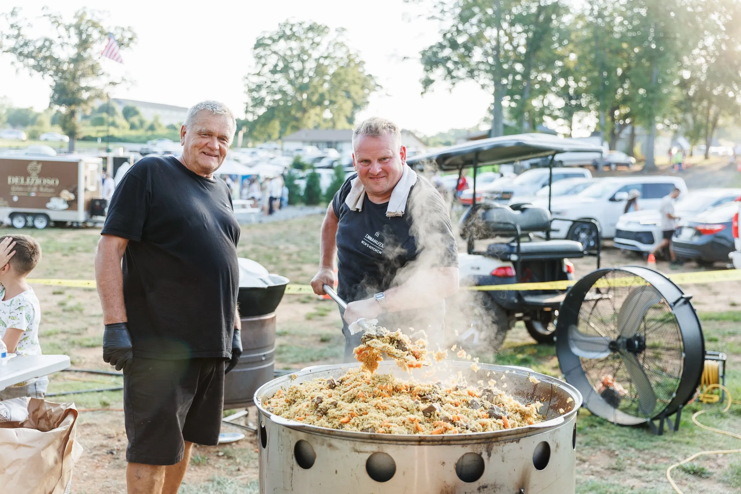 Two cooks stirring a giant pot of plov over an open fire at a community event