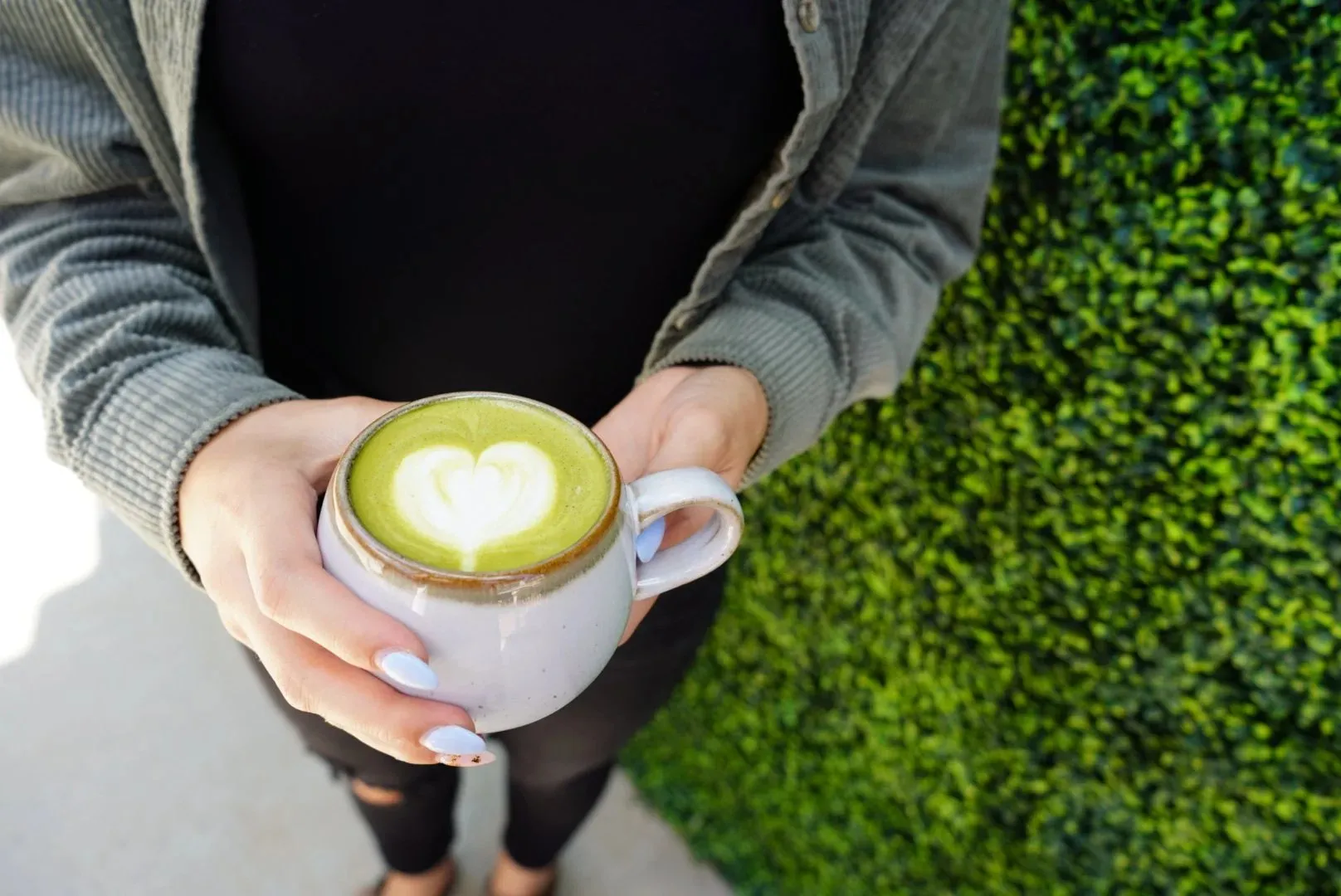 Hands holding a matcha latte with heart latte art beside a green hedge wall